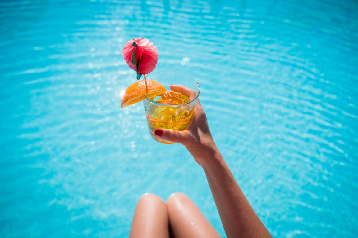 Una mano sosteniendo un cóctel con una guarnición de frutas sobre una piscina.