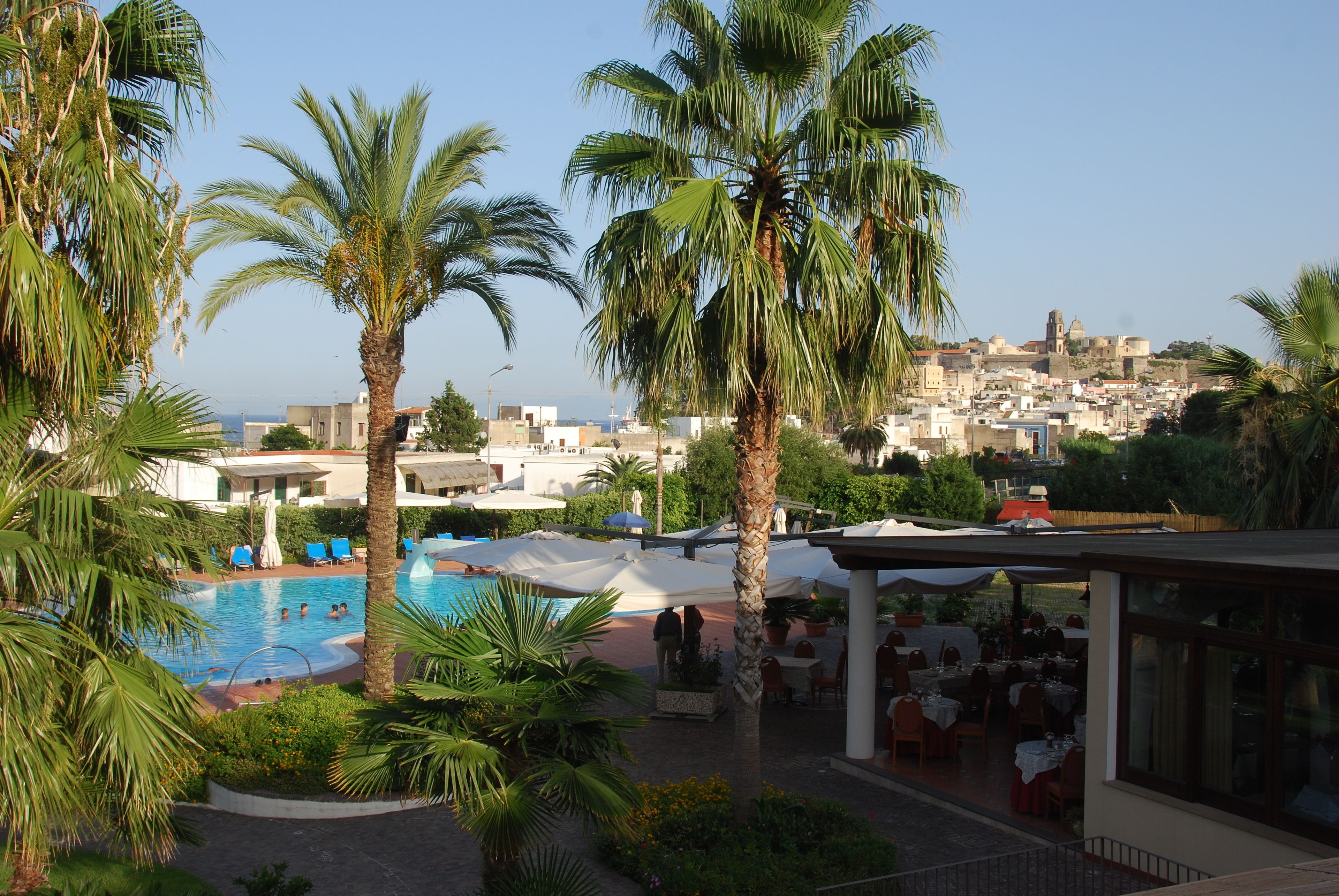 A resort pool area with palm trees, outdoor seating, and a view of a historic town in the background.