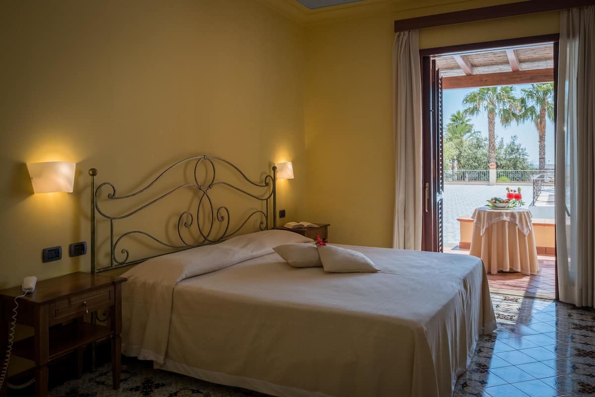 A neatly made bed with two bedside lamps, a small bedside table with a phone, and a view of a balcony with a table and chairs.