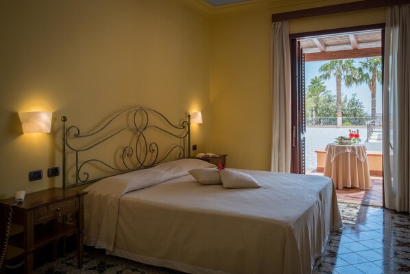 A neatly made bed with two bedside lamps, a small bedside table with a phone, and a view of a balcony with a table and chairs.