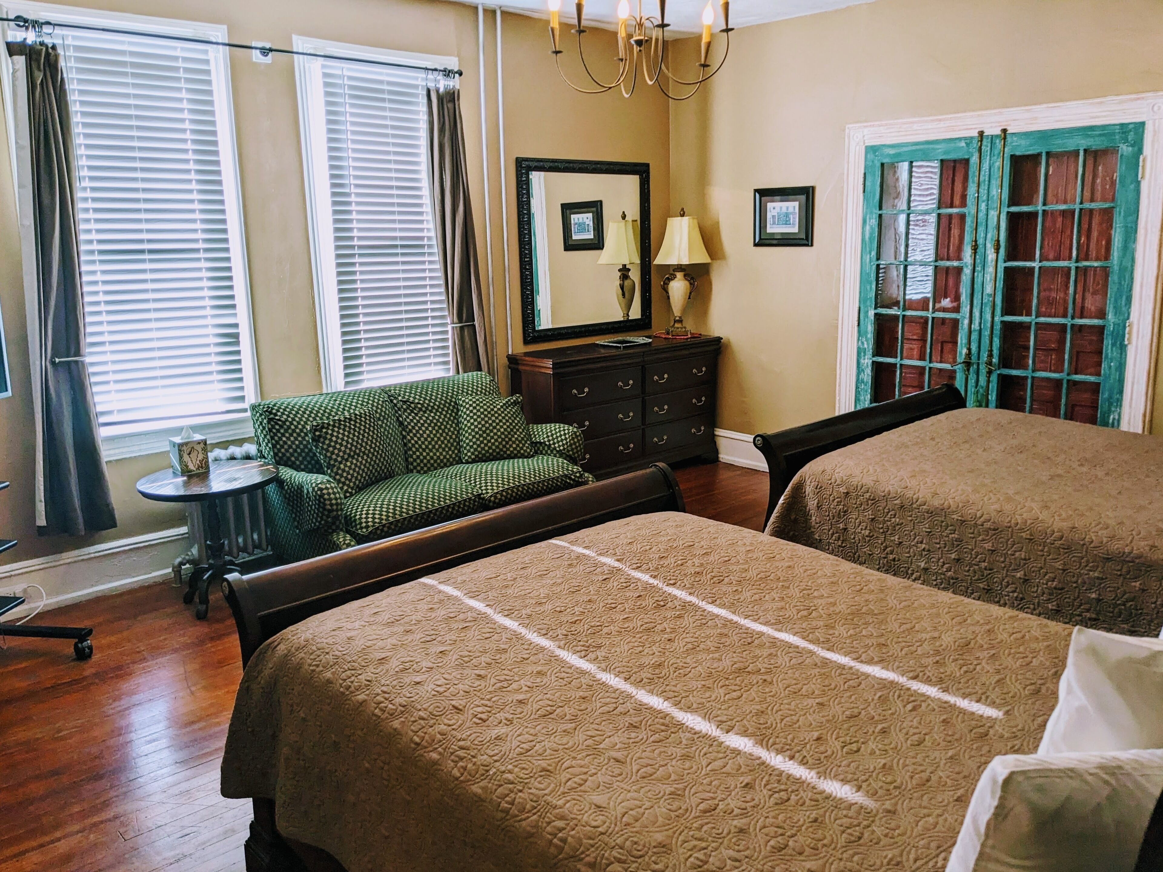 A room with two beds, a small table, a chair, a dresser, and a window with blinds.