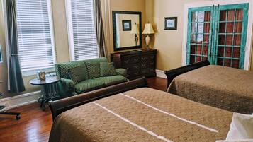 A room with two beds, a small table, a chair, a dresser, and a window with blinds.