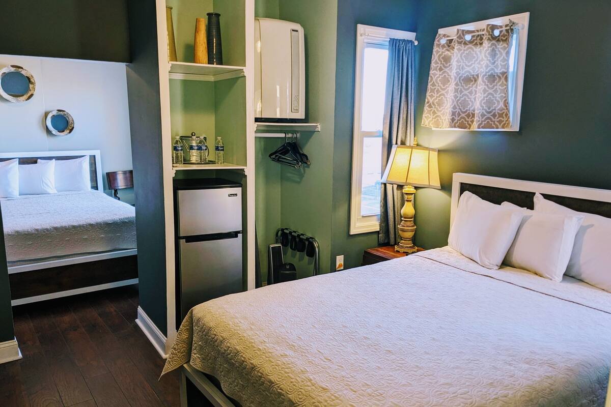 A hotel room with a bed, a refrigerator, a shelf with toiletries, a window with curtains, and a lamp.