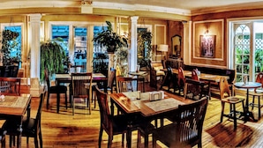 A spacious dining area with multiple wooden tables and chairs, large windows offering views of the outdoors, and various potted plants.