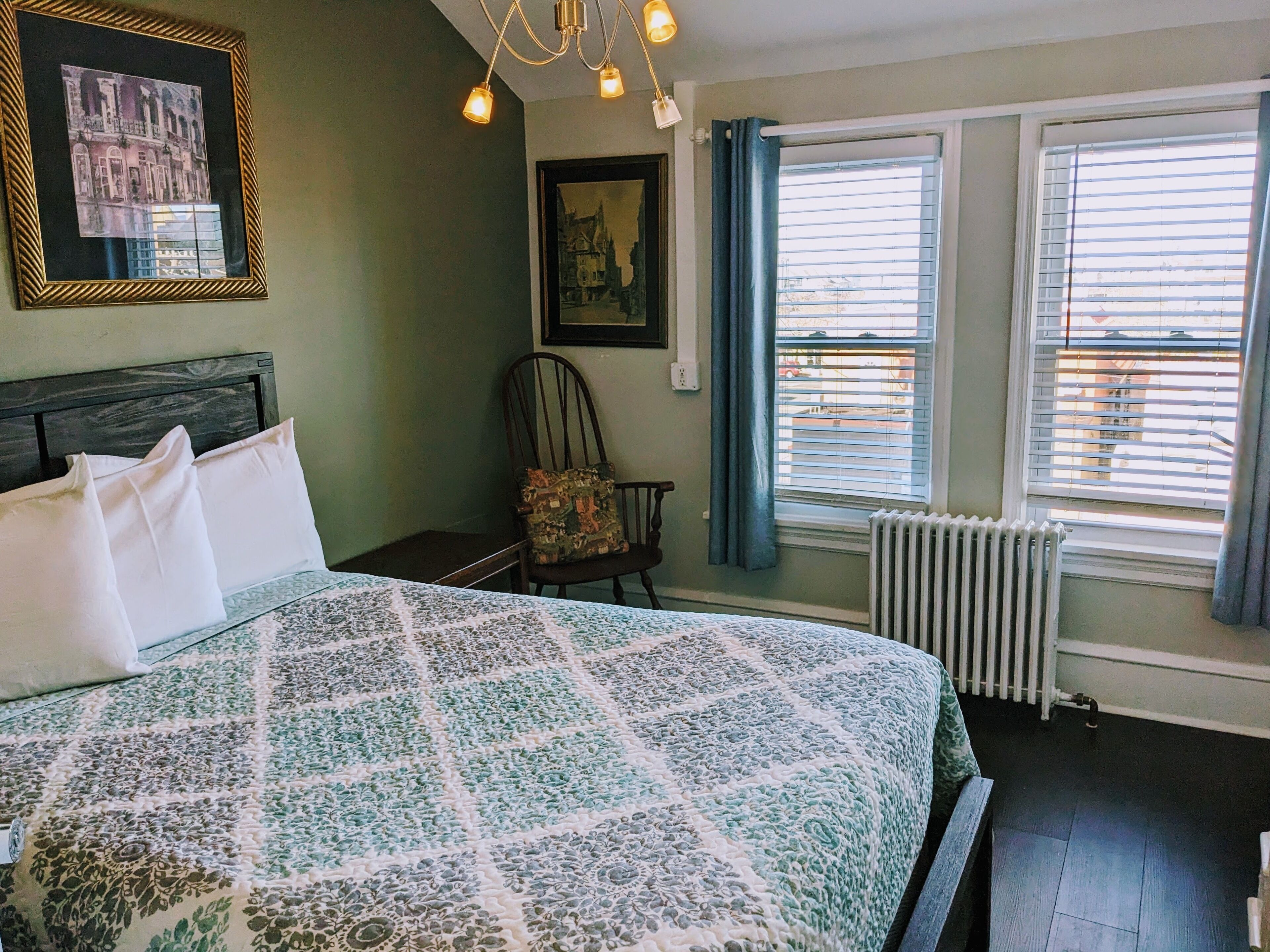 A neatly made bed with a patterned bedspread, a chair with a cushion, a framed picture on the wall, and a window with blinds.