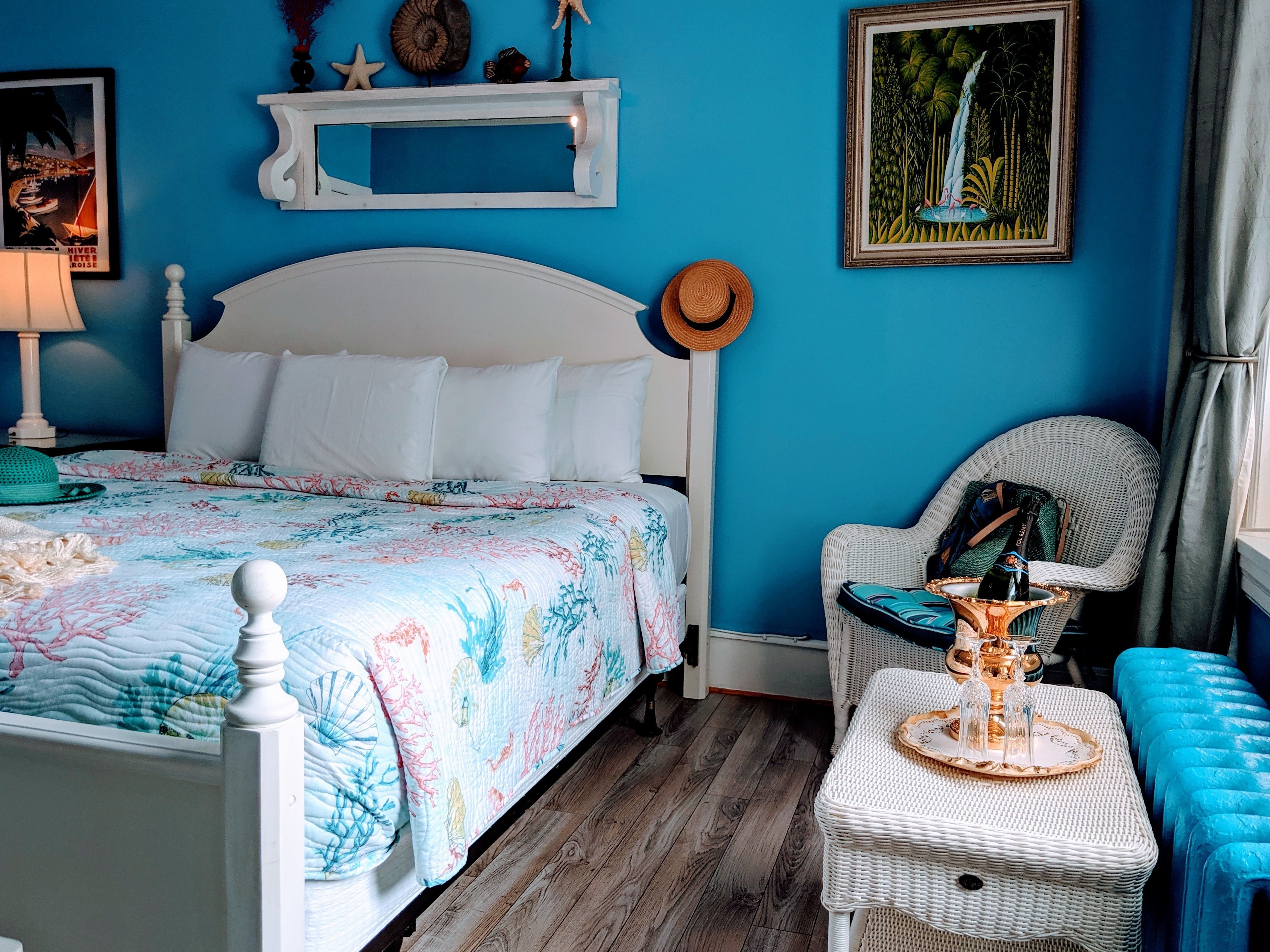 A bedroom with a bed, a wicker chair, a small table with a bottle, and a framed picture on the wall.