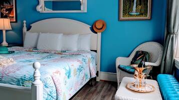 A bedroom with a bed, a wicker chair, a small table with a bottle, and a framed picture on the wall.