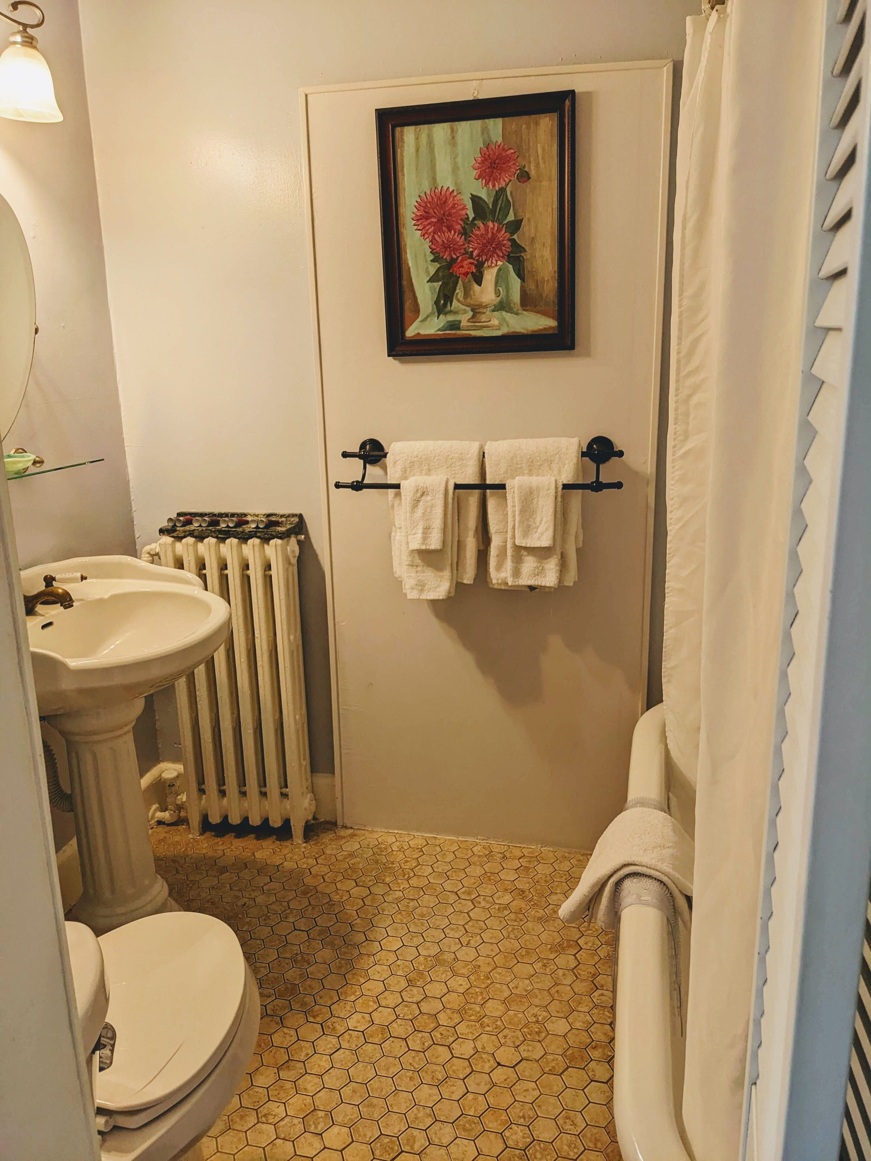 A bathroom with a toilet, sink, radiator, and a framed painting of flowers on the wall.