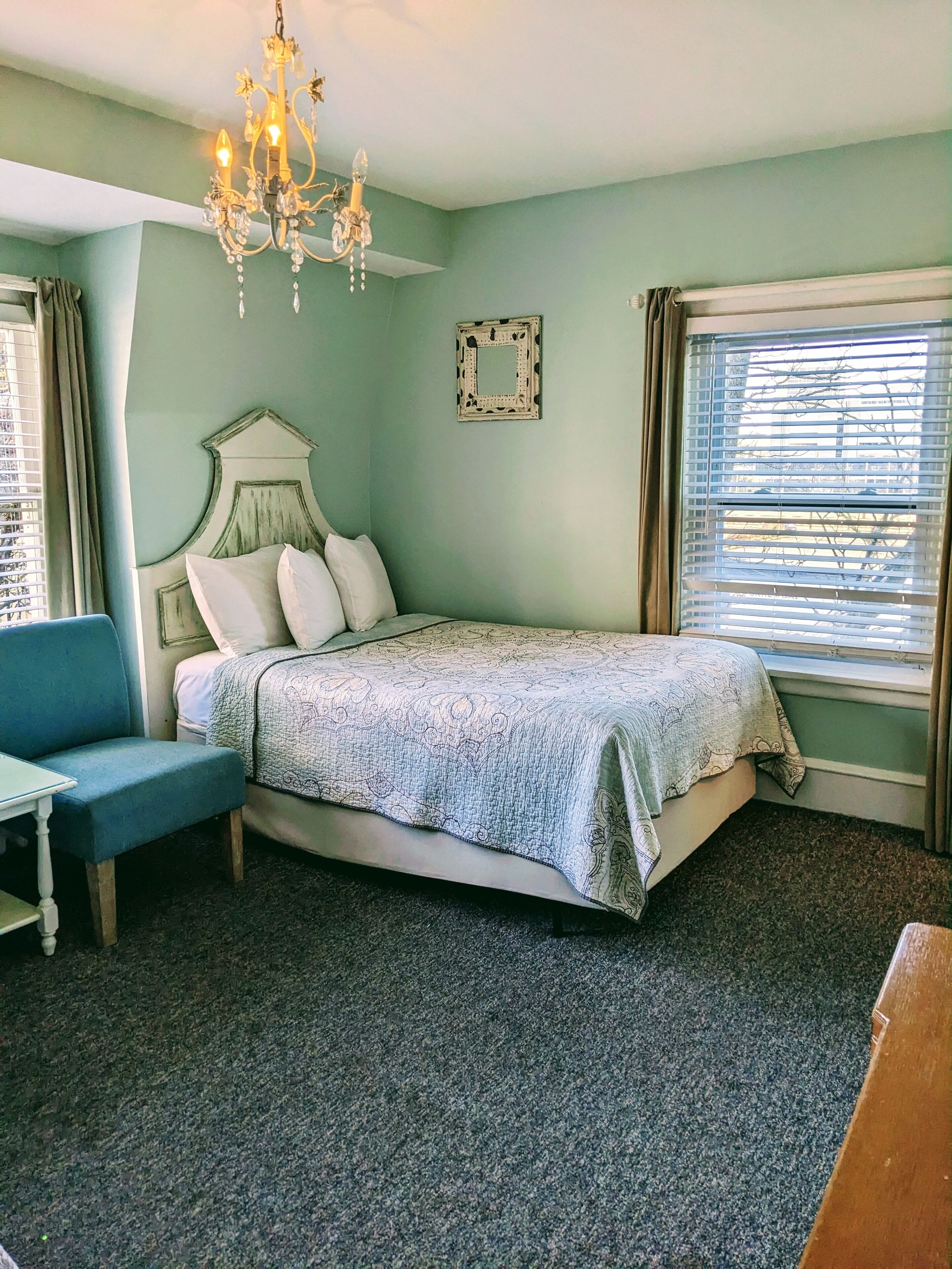 Un dormitorio con cama, una silla, candelabro, ventana con persianas y espejo adosado a la pared.