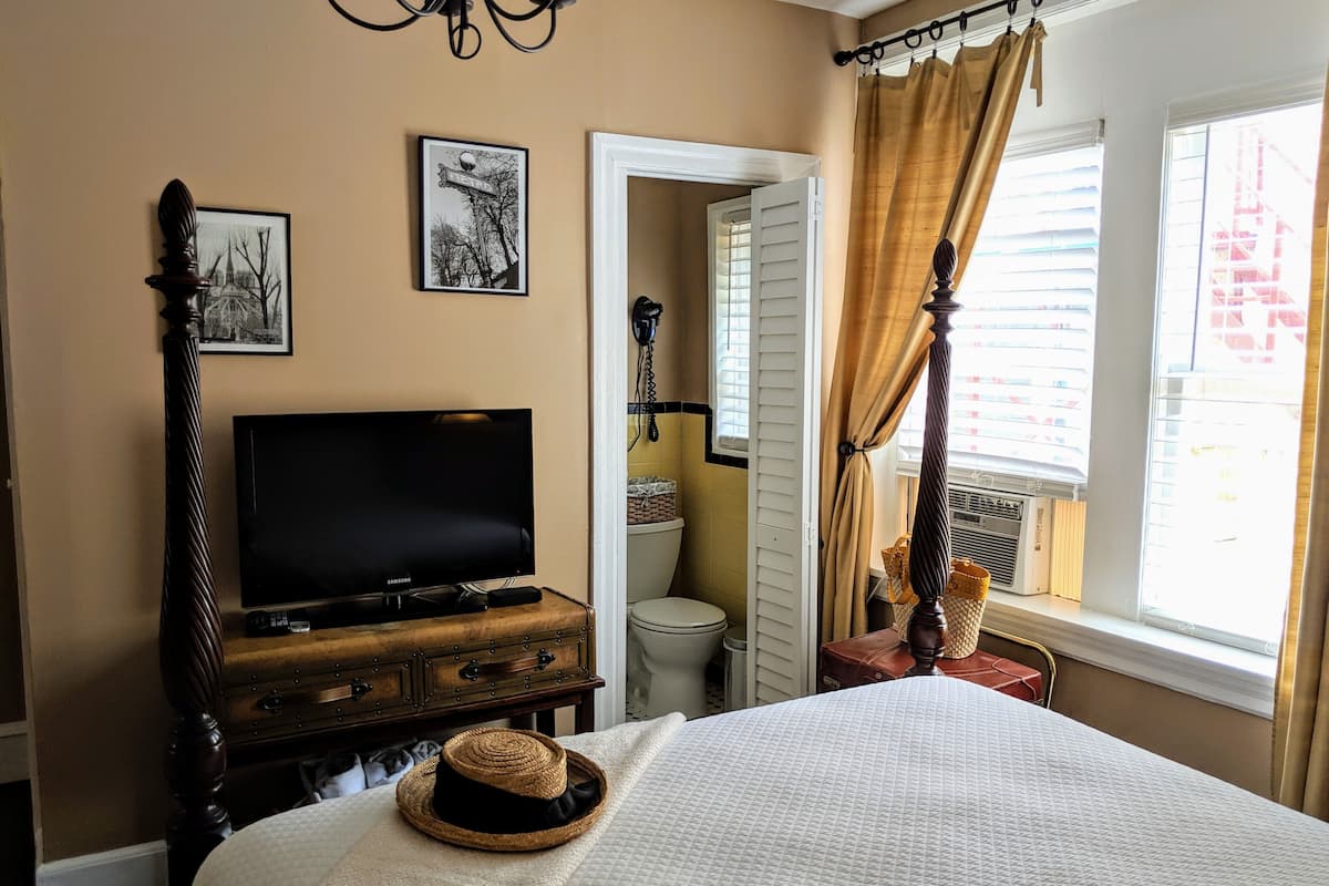 A bedroom with a bed, a television on a wooden dresser, a door leading to a bathroom, and a window with curtains.