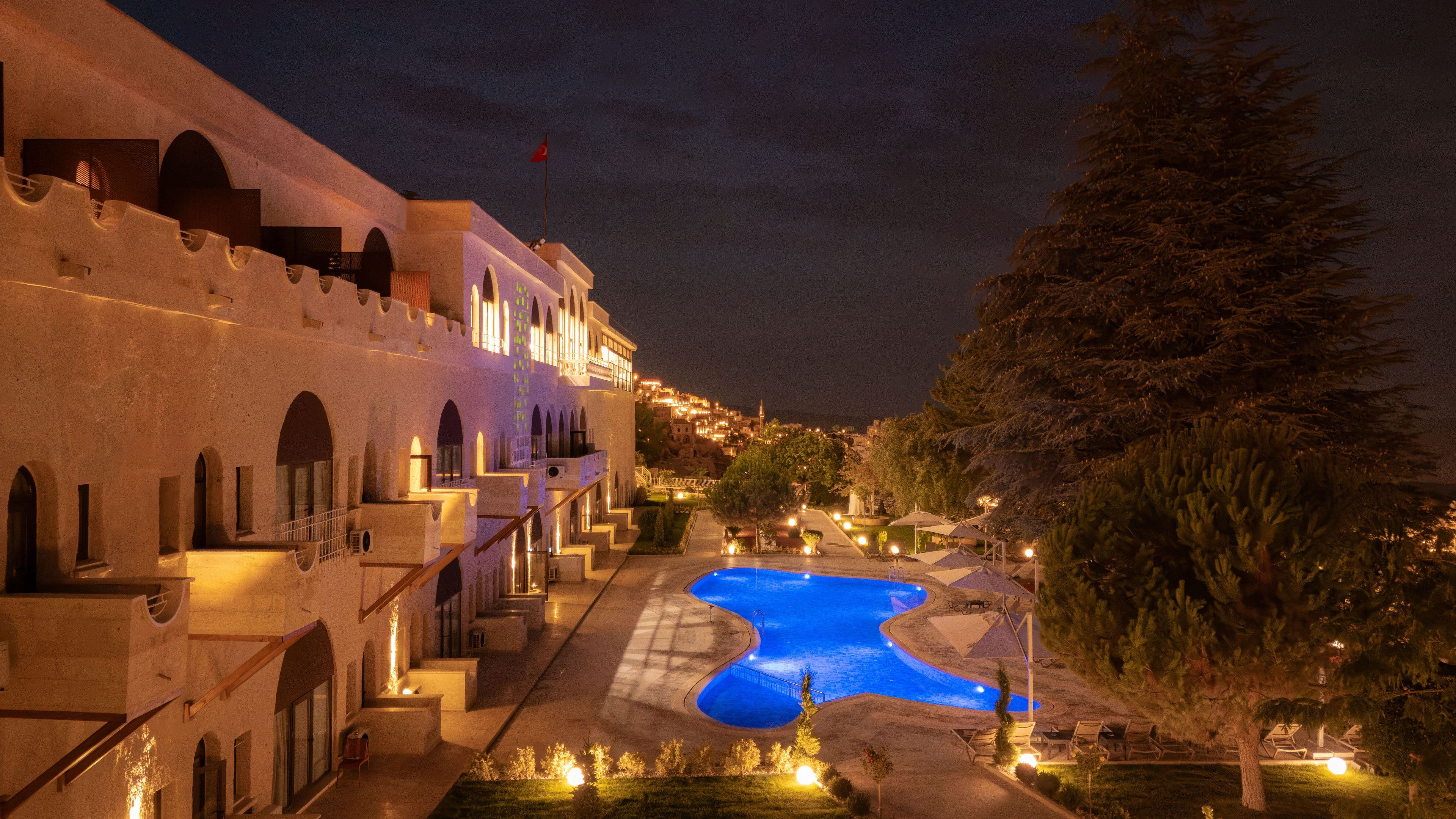 A nighttime view of a hotel with a swimming pool, arched balconies, and a view of the city in the background.