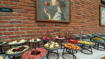Disposição de buffet com diversos alimentos em bandejas, em frente a uma parede de tijolos com um quadro emoldurado.