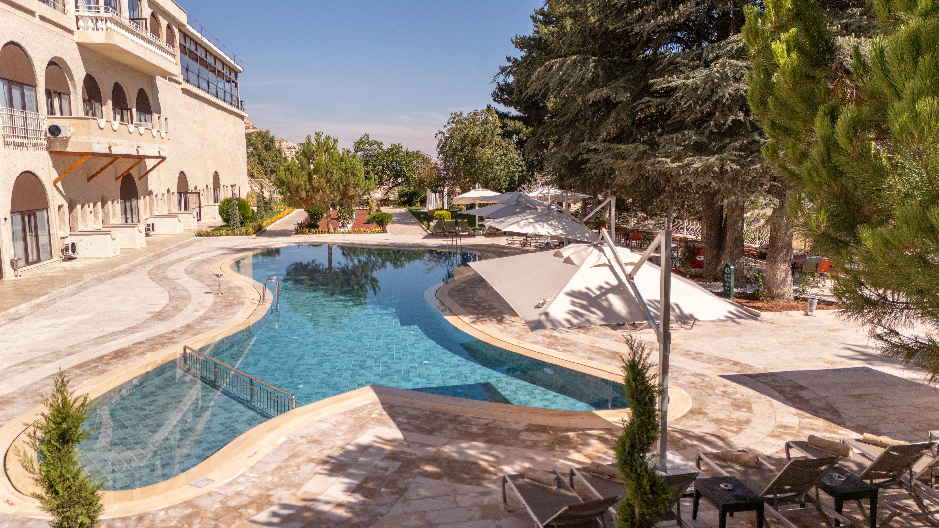 Una piscina con diseño curvo, rodeada de sillones de descanso y sombrillas, con un edificio al fondo.