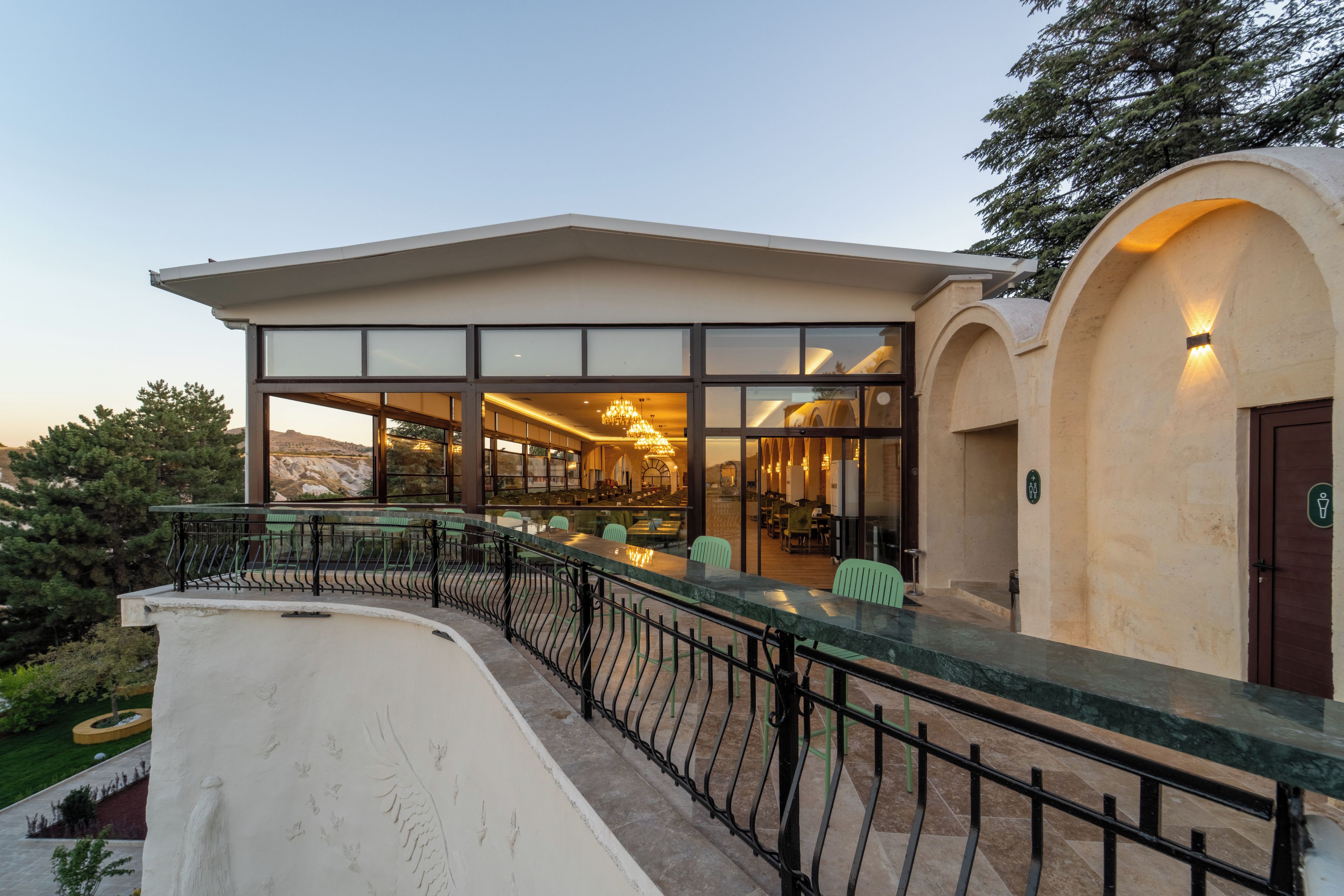 A modern building with a covered outdoor seating area, large windows, and a view of the landscape.