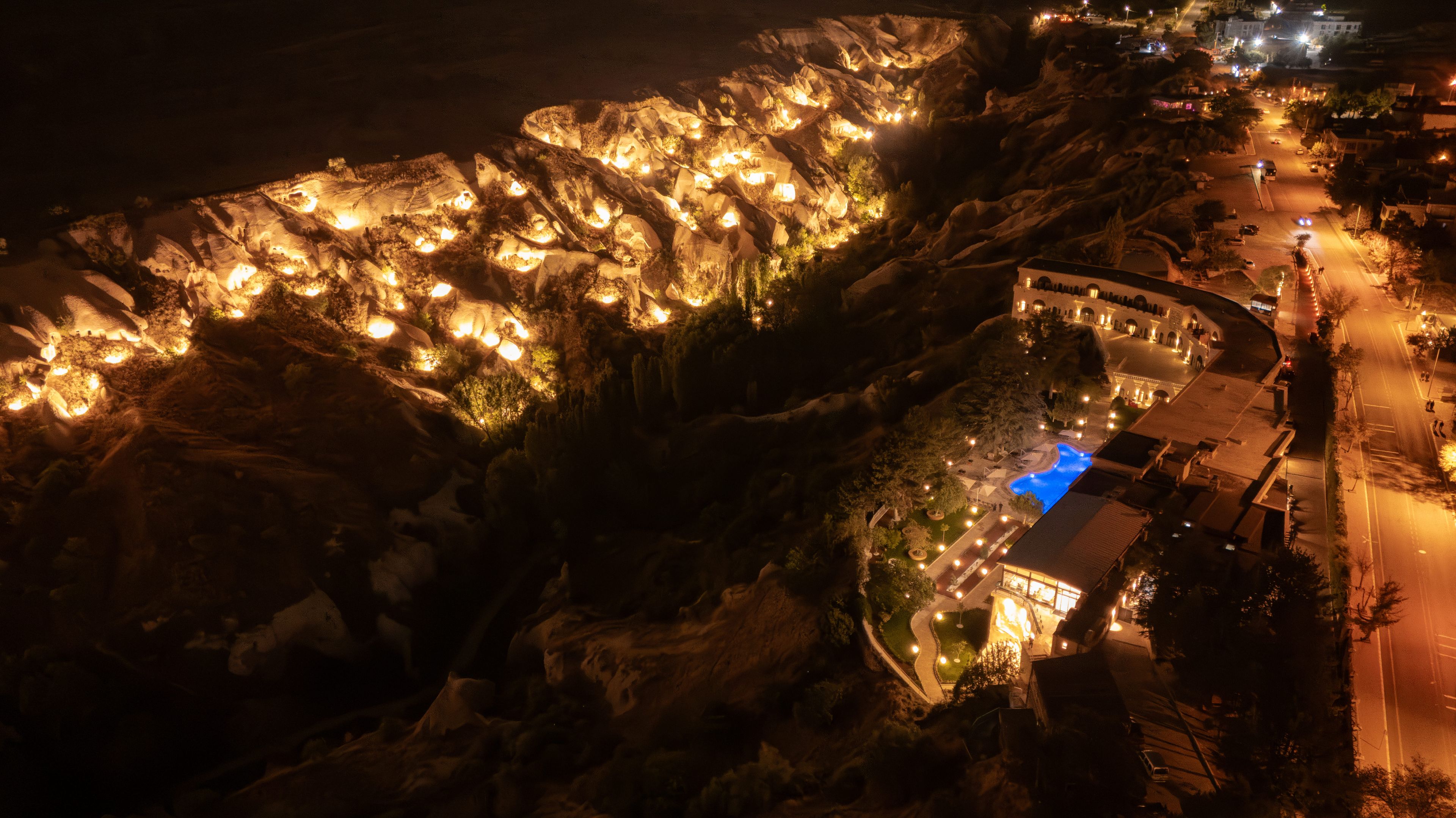 Vista aérea nocturna de una propiedad costera con formaciones rocosas iluminadas, una piscina y un camino que lleva a un edificio.
