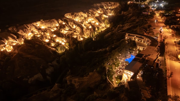Une vue aérienne nocturne d’une propriété située sur la côte, avec des formations rocheuses illuminées, une piscine et une route menant à un bâtiment.