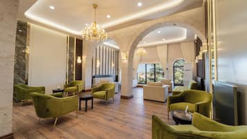 A spacious lobby with green upholstered chairs, a grand chandelier, and a view through an arched doorway to a balcony.