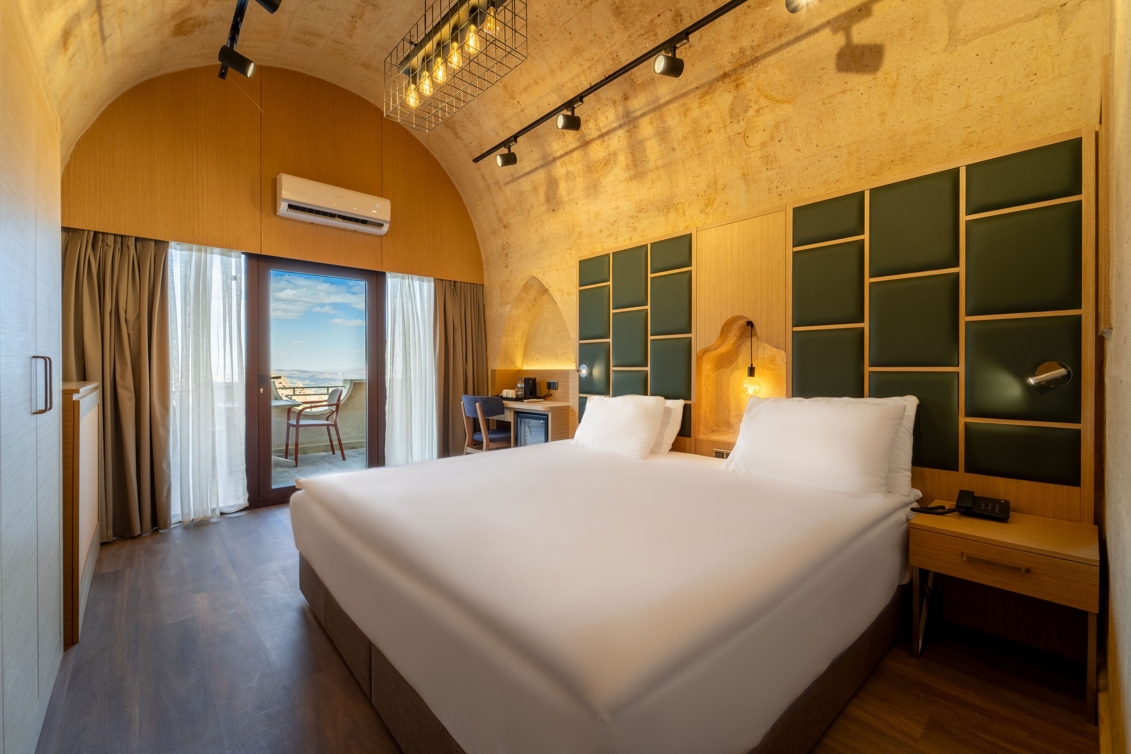 Habitación de hotel con una cama grande, un escritorio con silla, vista a través de un ventanal y un diseño moderno en el techo.