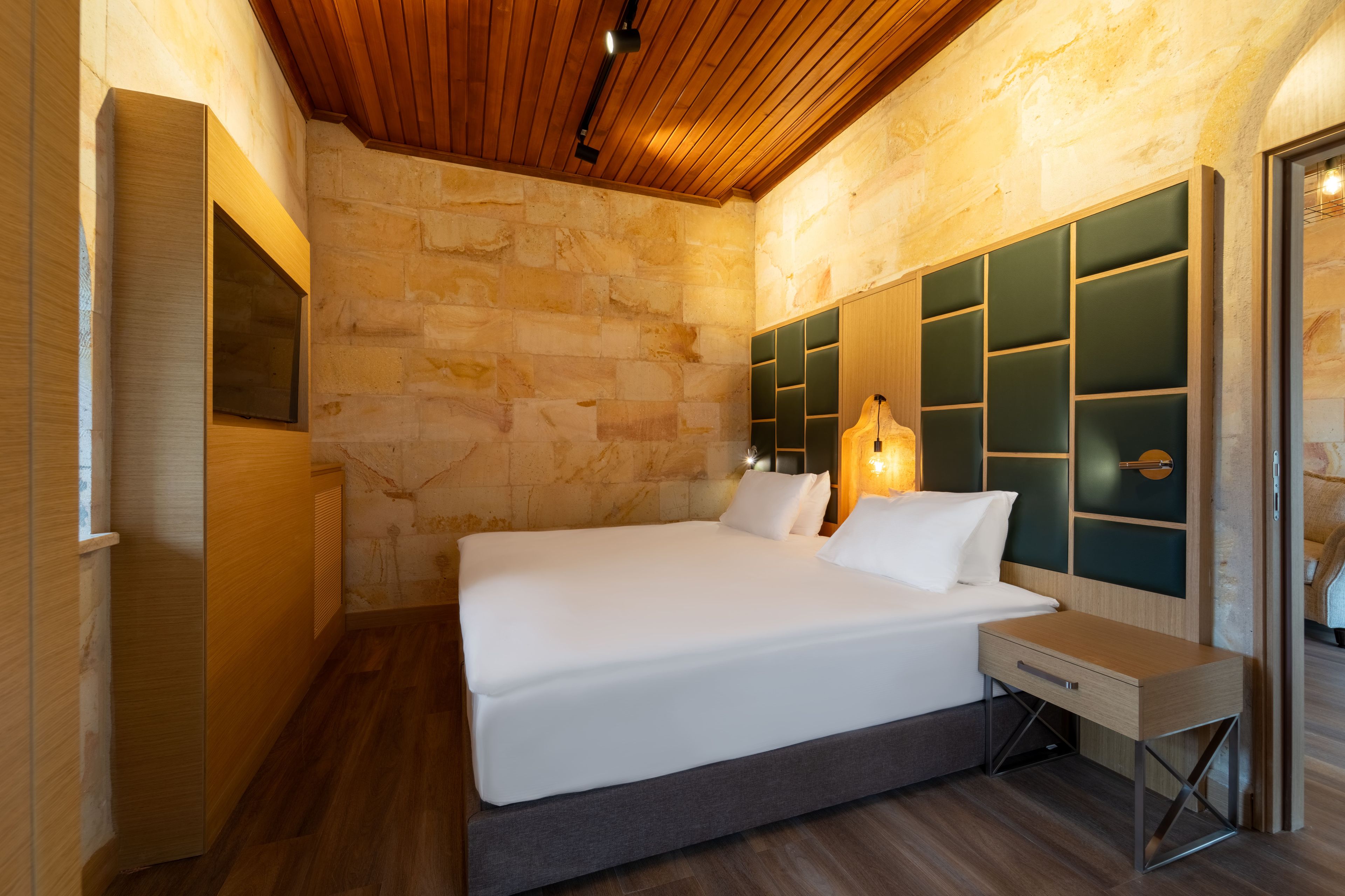 A modern bedroom with a wooden ceiling, stone walls, a large bed with a headboard, bedside tables, and a wall-mounted TV.