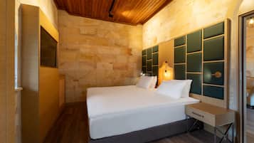 A modern bedroom with a wooden ceiling, stone walls, a large bed with a headboard, bedside tables, and a wall-mounted TV.