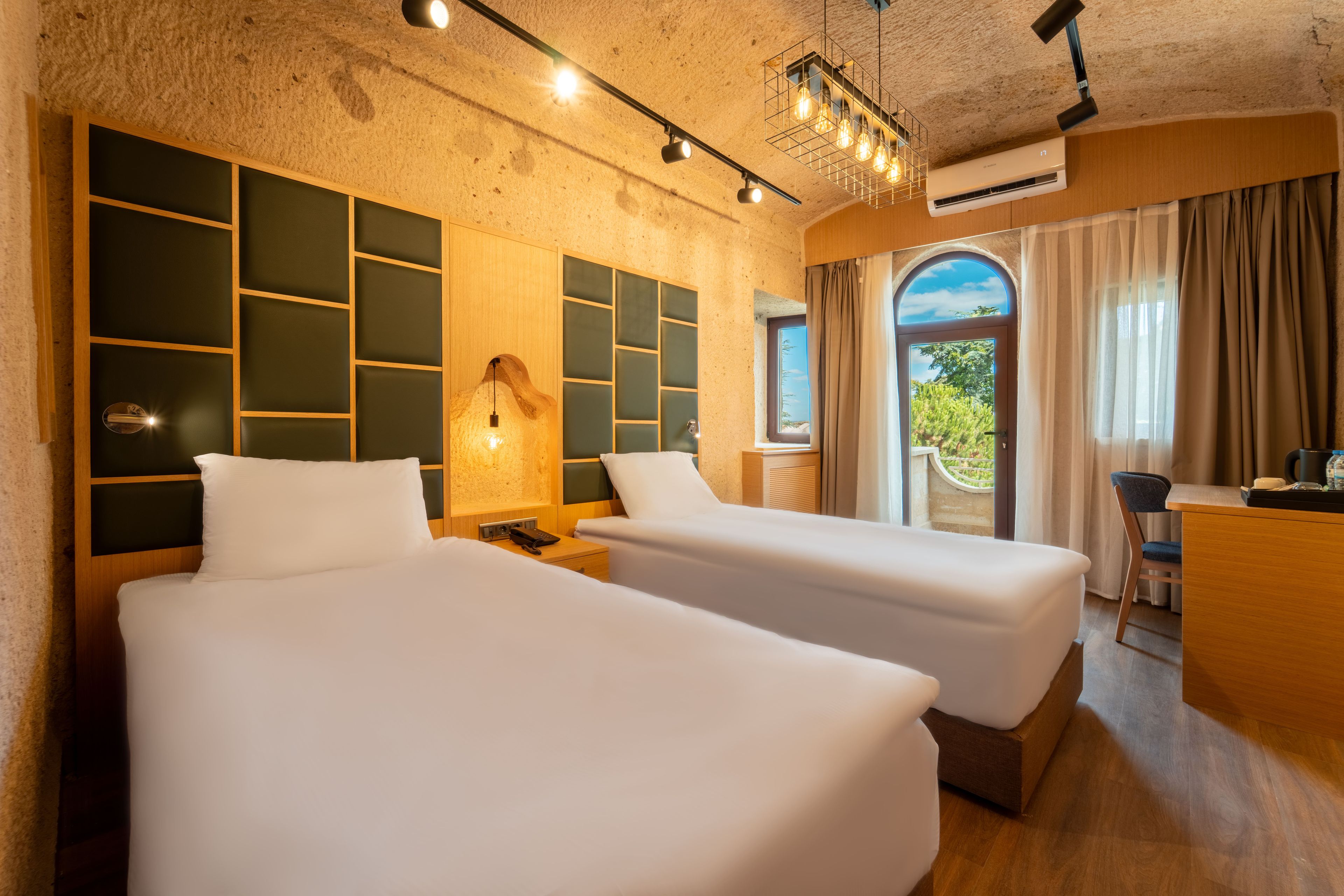 Quarto de hotel com duas camas, uma escrivaninha, uma luminária e vista para o verde.