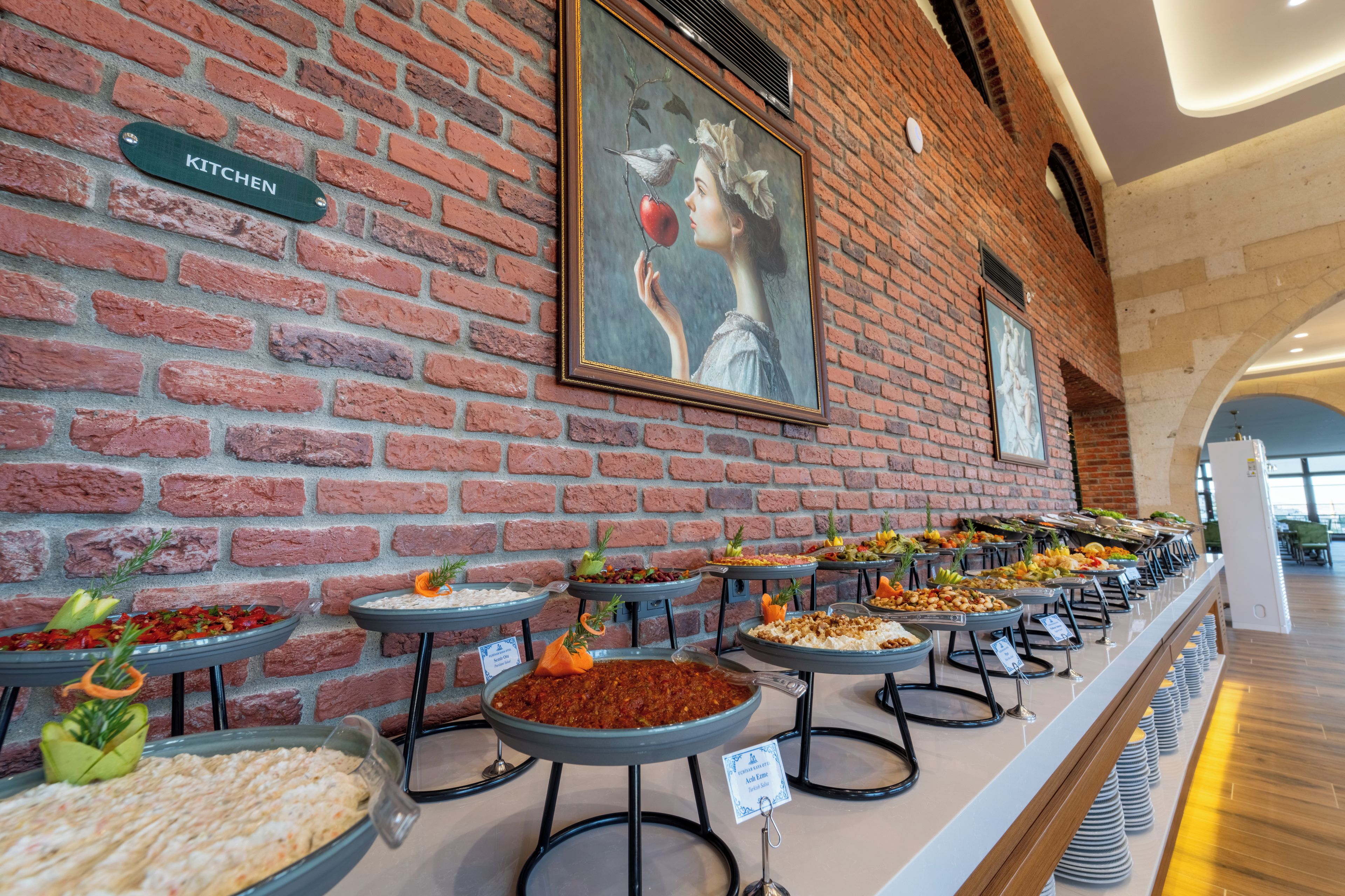 Zona de bufé con diferentes estaciones de comida, pared de ladrillo con un cartel que dice 'KITCHEN' y un cuadro enmarcado en la parte superior.