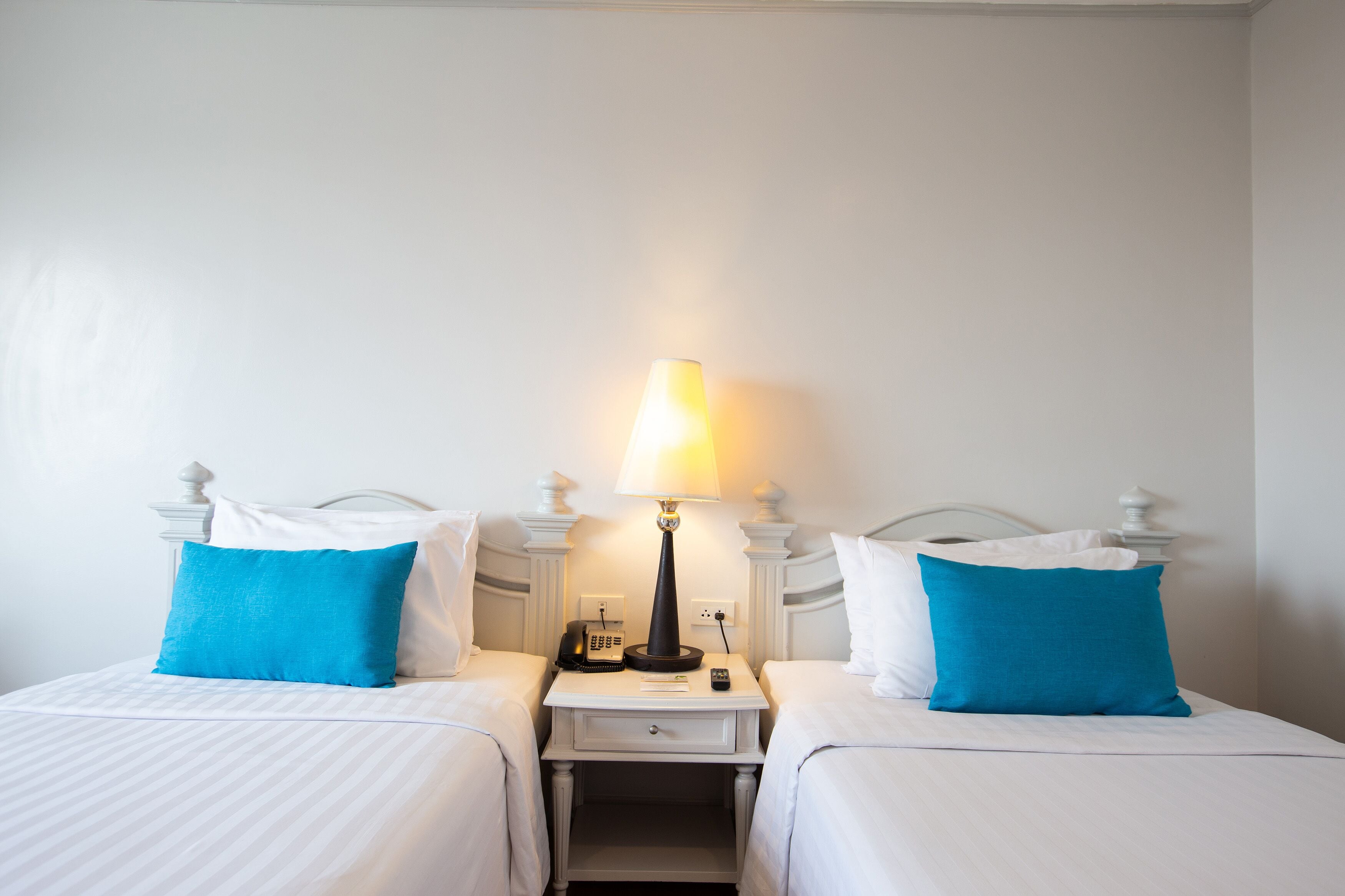 Une chambre d’hôtel avec deux lits, chacun recouvert d’une courtepointe blanche et agrémenté d’un oreiller bleu. Une table de chevet se trouve entre les deux lits, sur laquelle on peut trouver une lampe, un téléphone et une télécommande.