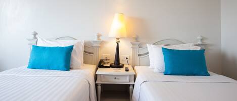 Une chambre d’hôtel avec deux lits, chacun recouvert d’une courtepointe blanche et agrémenté d’un oreiller bleu. Une table de chevet se trouve entre les deux lits, sur laquelle on peut trouver une lampe, un téléphone et une télécommande.