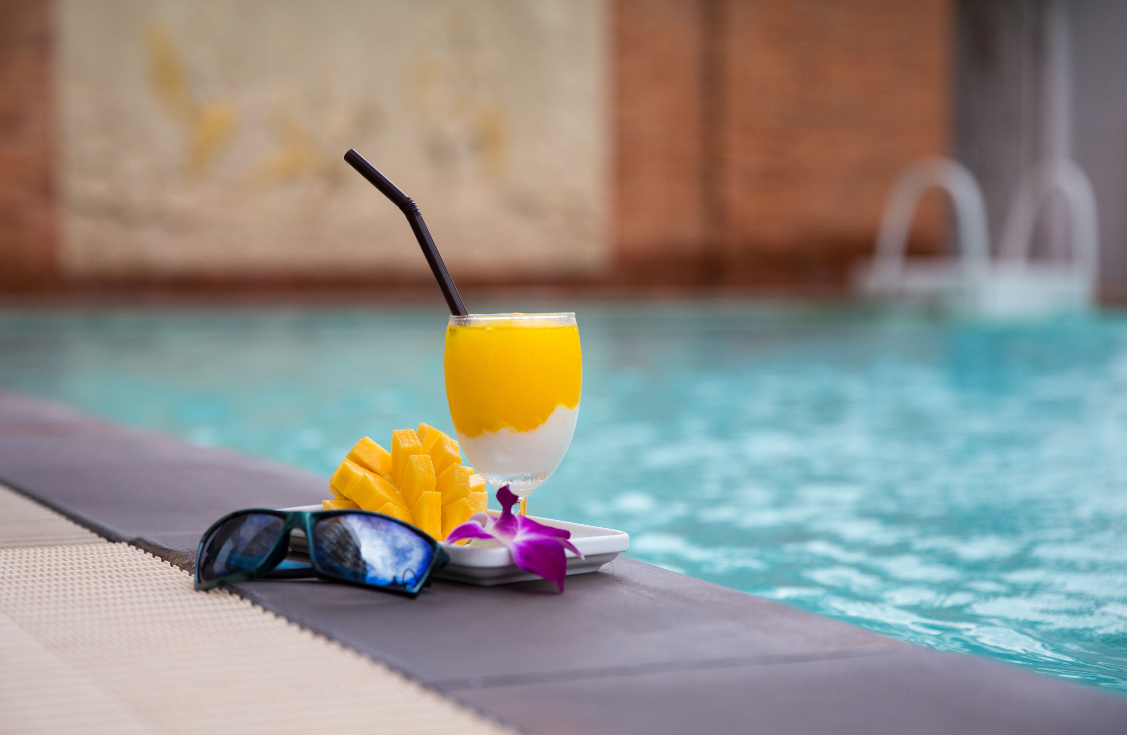Un vaso de jugo de naranja con una pajita, un par de gafas de sol y un mango sobre una mesa junto a la piscina.