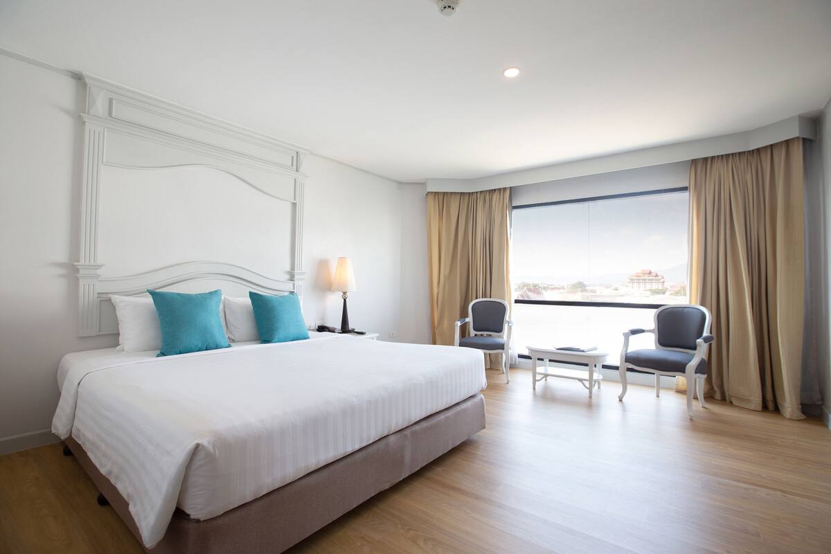 A hotel room with a large bed, two chairs, a small table, and a view of the city through large windows.