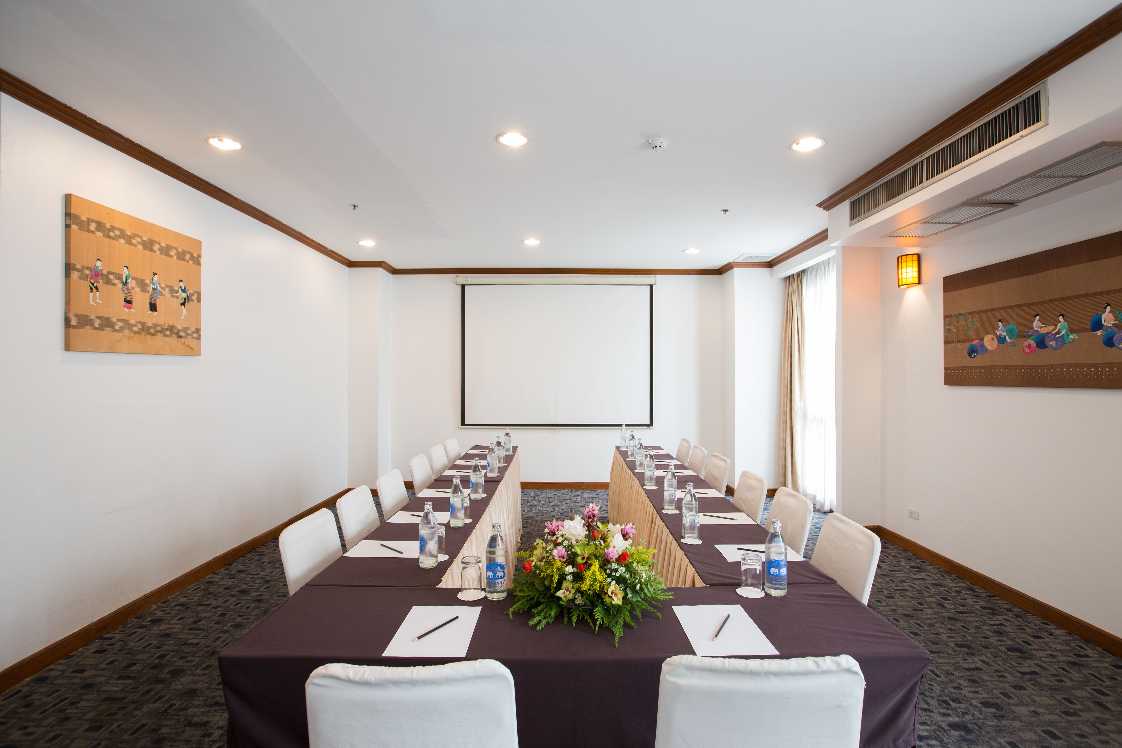 Un salón de conferencias con una larga mesa, sillas, una pantalla de proyector y centros de mesa florales.