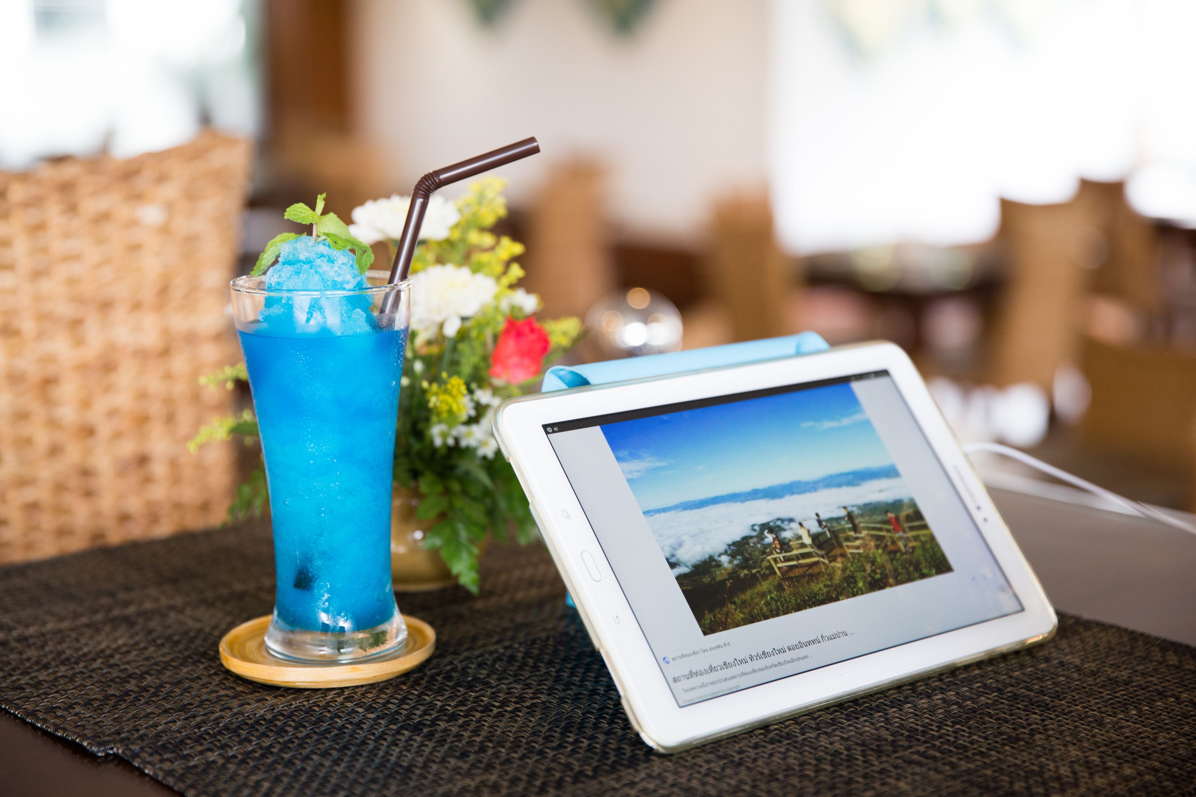A tablet displaying a scenic landscape, a glass of blue beverage with a straw, and a small vase with flowers on a table.