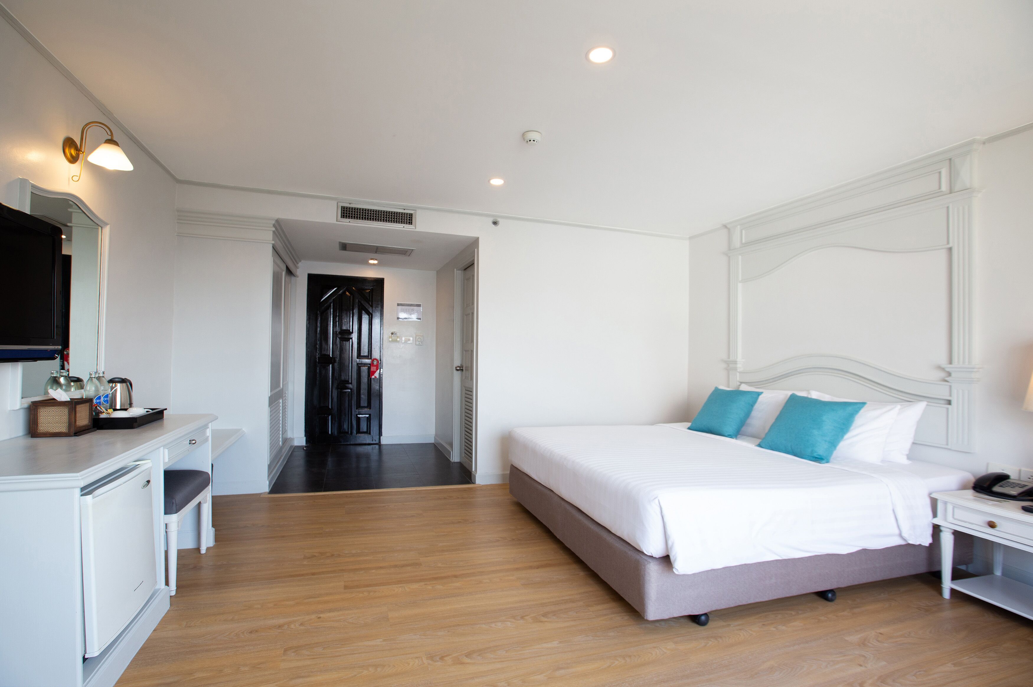 A hotel room with a bed, bedside table, and a desk with a chair. There is a mirror on the wall and a door leading to another room.