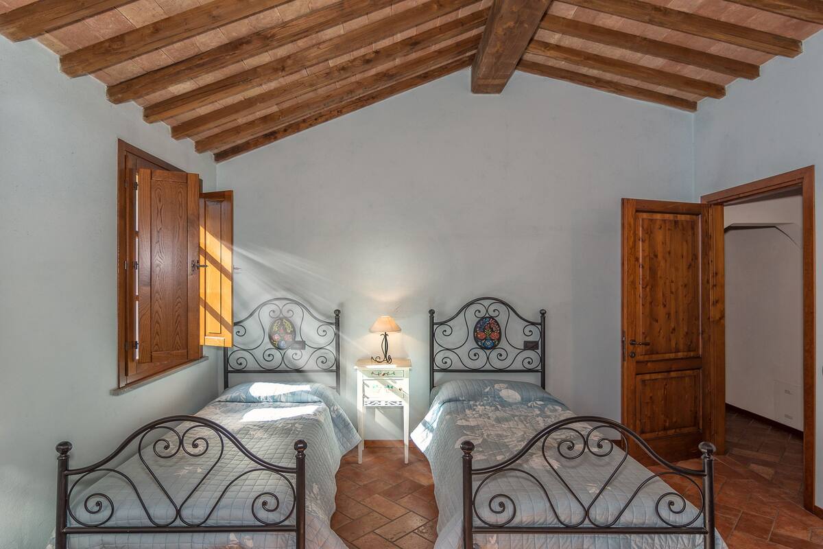 A bedroom with two beds, wooden ceiling beams, a nightstand with a lamp, and a wooden door.