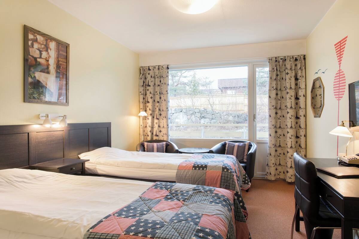 A hotel room with two beds, a desk, a chair, a window with curtains, and a painting on the wall.
