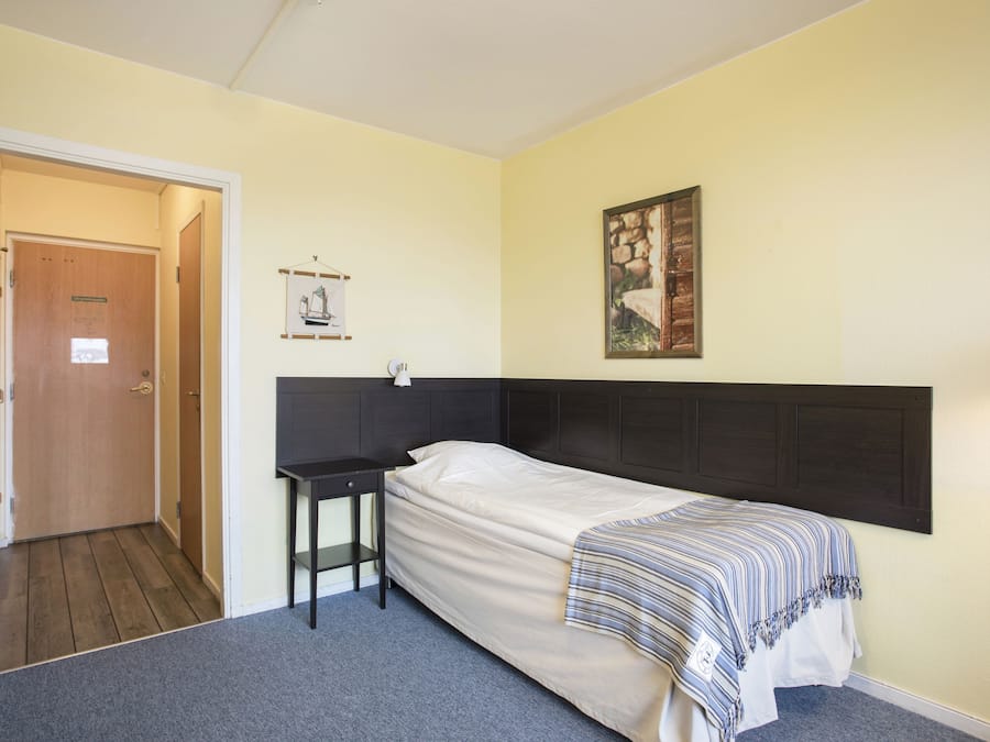 Una cama individual con cabecera oscura, una mesita de noche, un cuadro en la pared y una puerta que da a otra habitación.