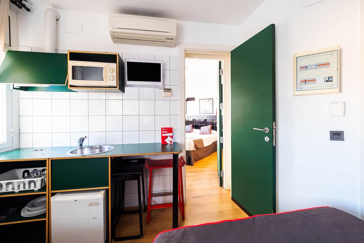 Una cocina compacta con microondas, lavamanos y un refrigerador pequeño. Hay una puerta verde que da a un dormitorio con una cama y una mesita de noche.