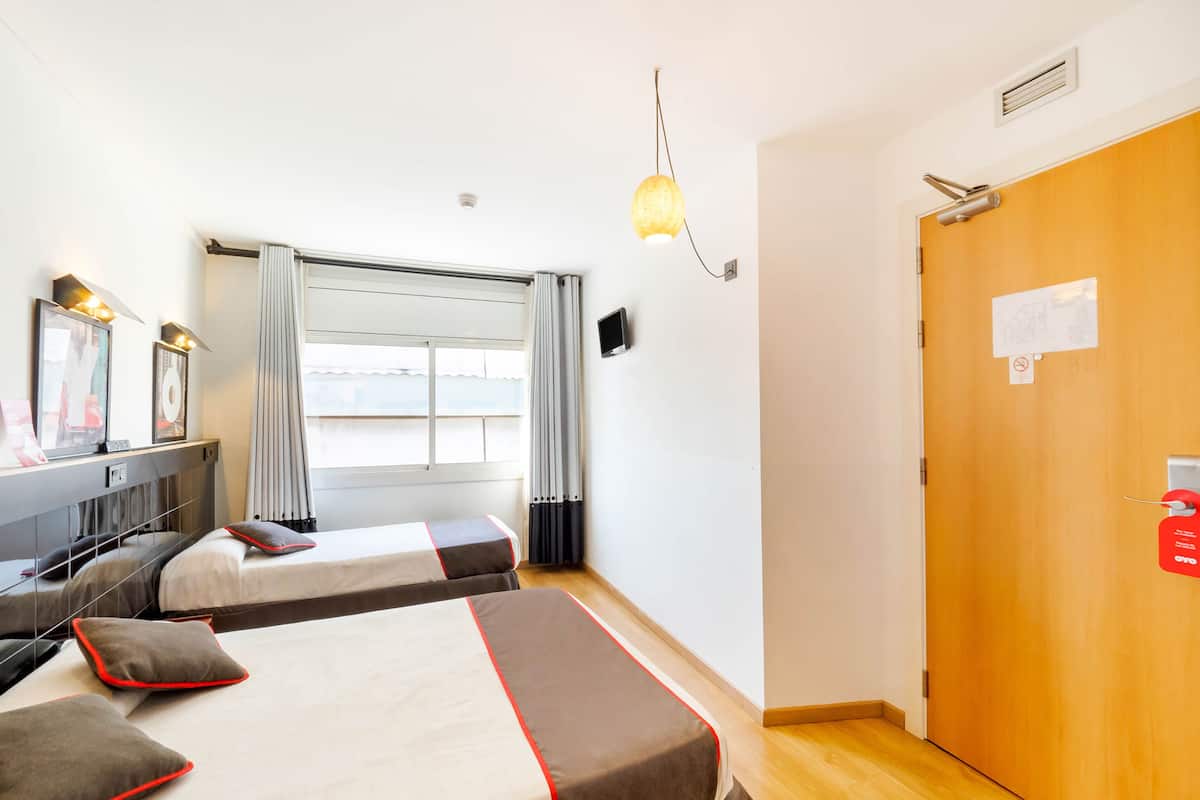 Una habitación de hotel con dos camas, un escritorio con computadora, una ventana con cortinas y una puerta de madera.