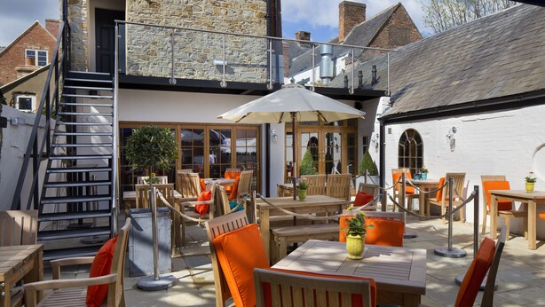An outdoor patio with wooden tables and chairs, a staircase, and stone walls. - The George Townhouse (Shipston on Stour)