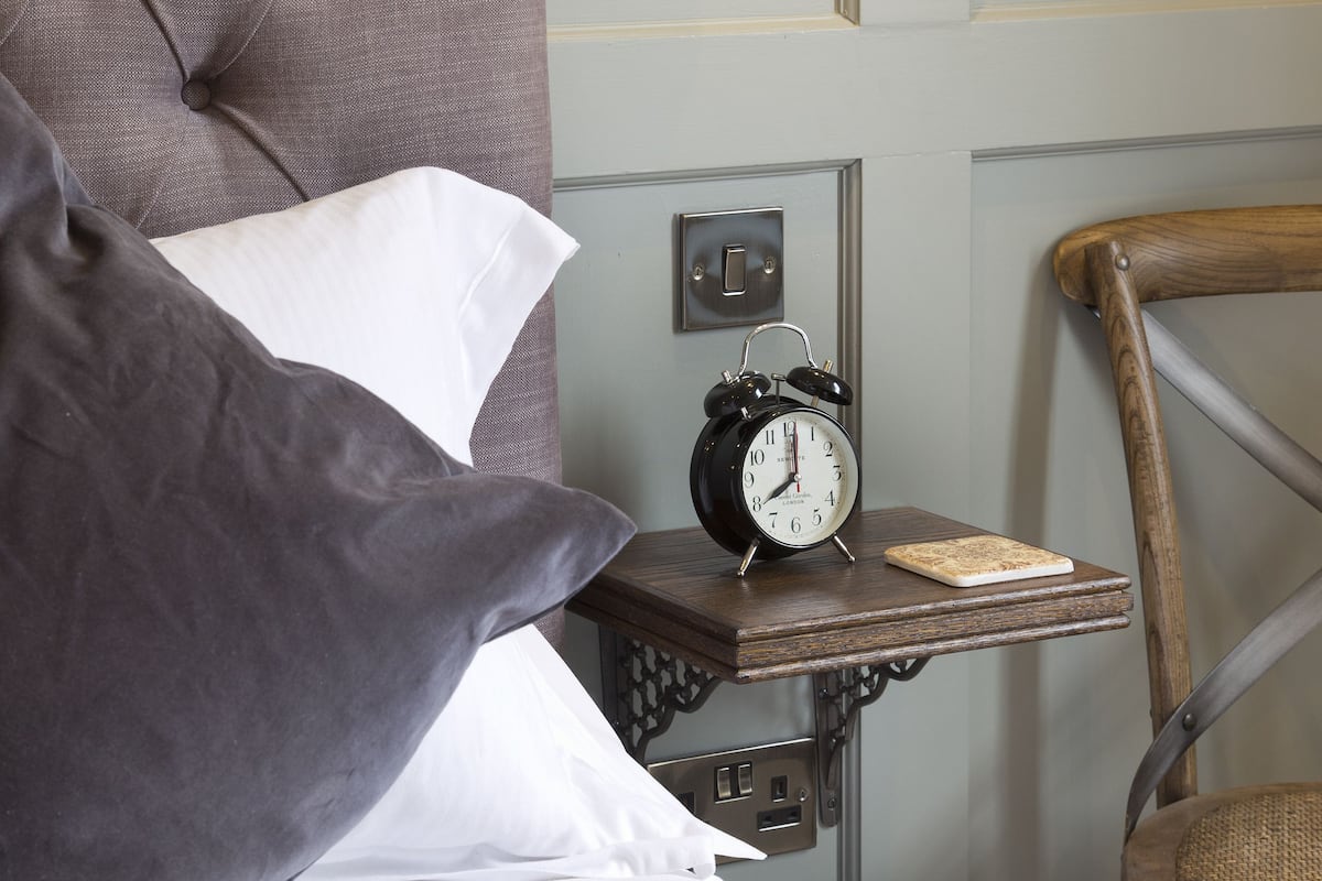 A neatly made bed with a grey headboard, a bedside table with an alarm clock, and a wall-mounted light fixture.
