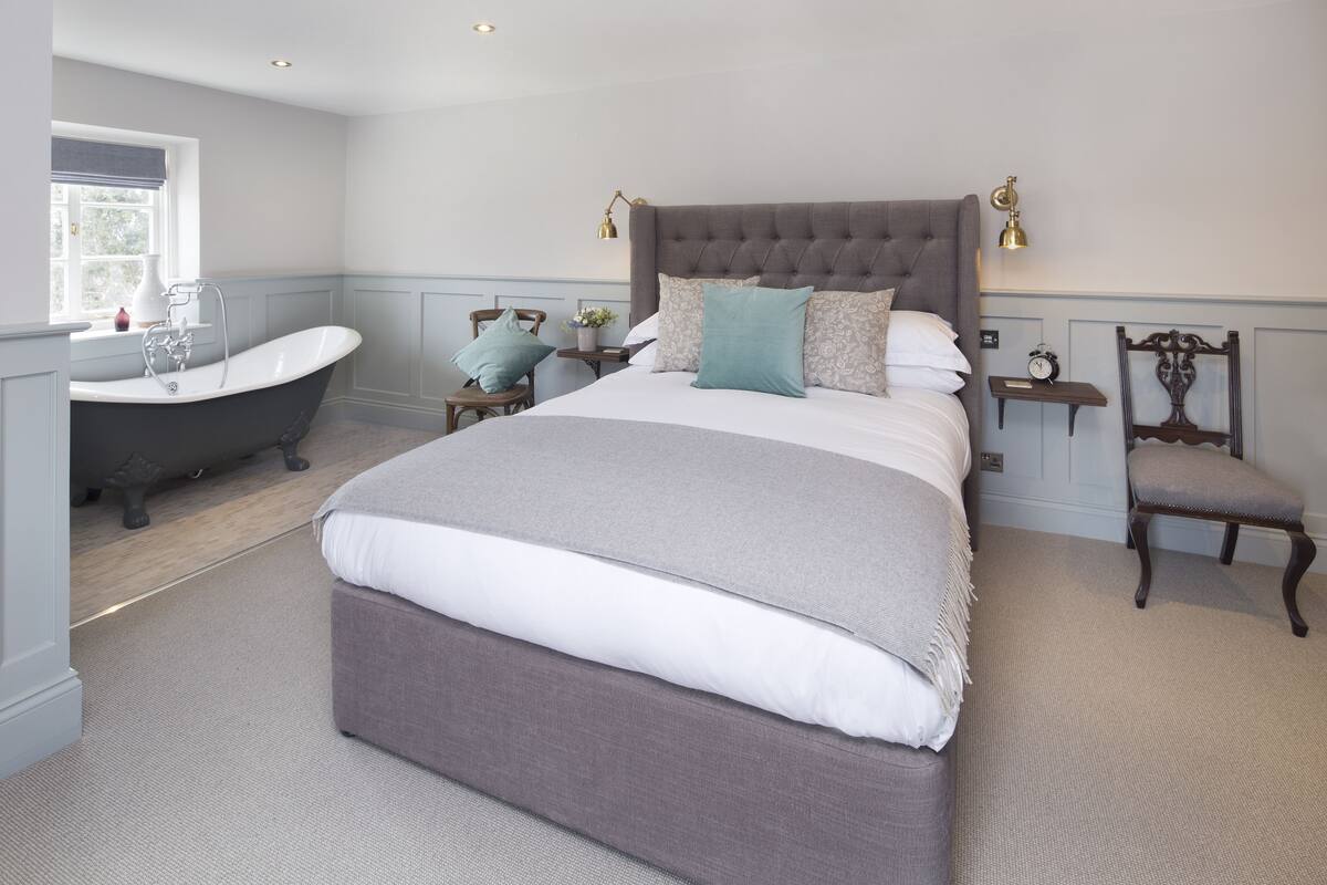 A bedroom with a large bed, a classic bathtub, a bedside table with a lamp, and a chair.