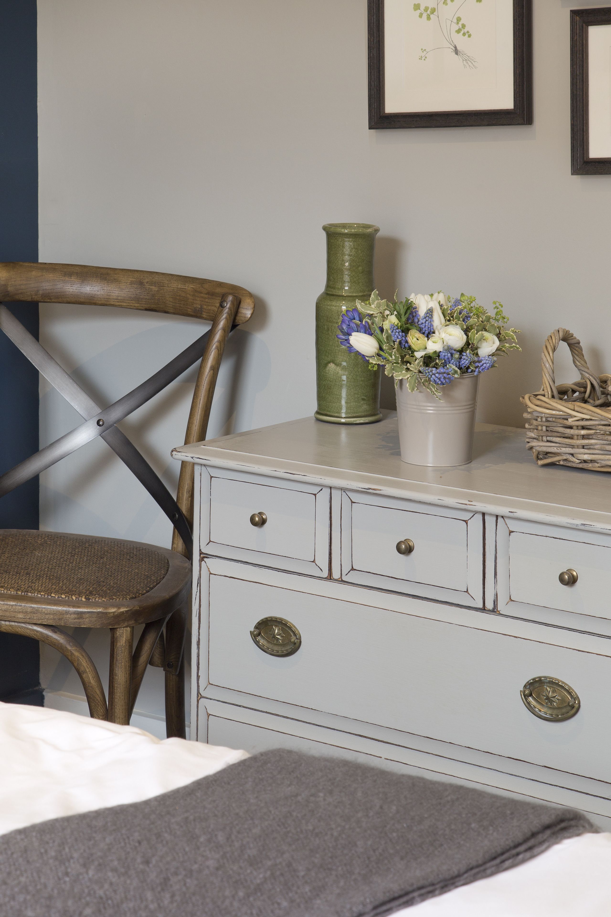 A room with a wooden chair, a white dresser with drawers, a green vase with flowers, and a framed picture on the wall.