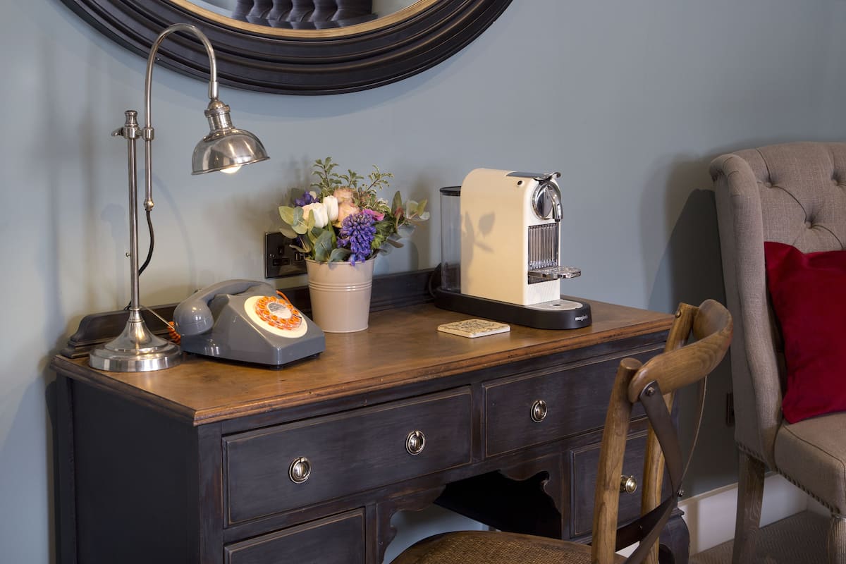 A wooden desk with a rotary phone, a coffee maker, a lamp, and a vase with flowers.