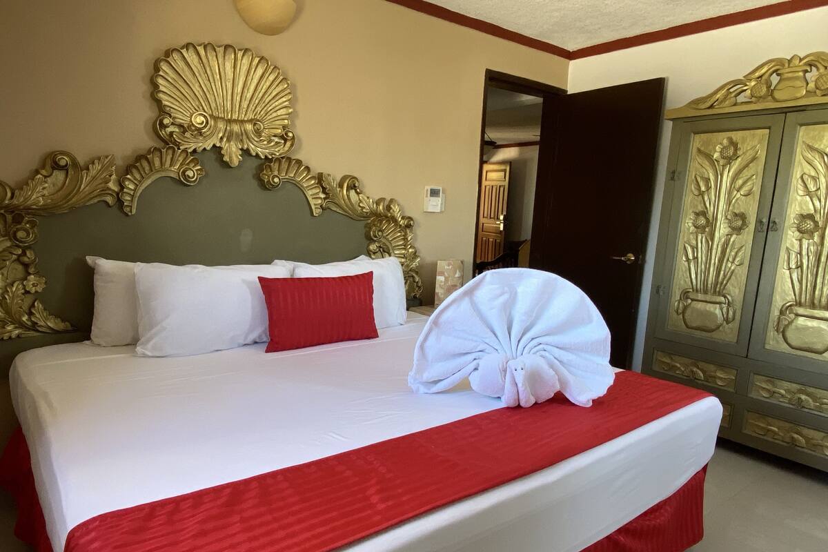 A hotel room with a large bed, a red and white bedspread, a red pillow, and a gold ornate headboard.