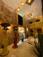 Un vestíbulo amplio con techo alto, paredes de piedra y lámparas modernas. Hay plantas en macetas y una zona de estar con una mesa y sillas.