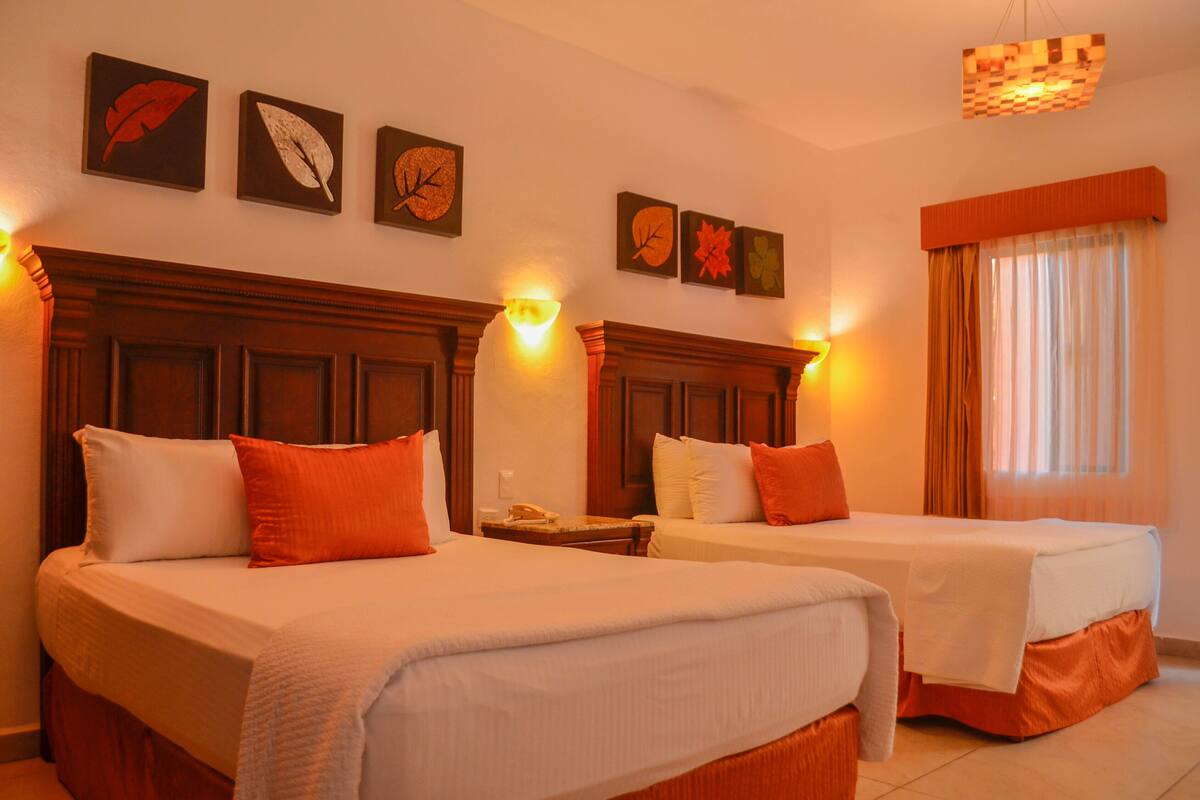 A hotel room with two beds, each with white bedding and orange pillows, a wooden headboard, and a nightstand with a lamp.
