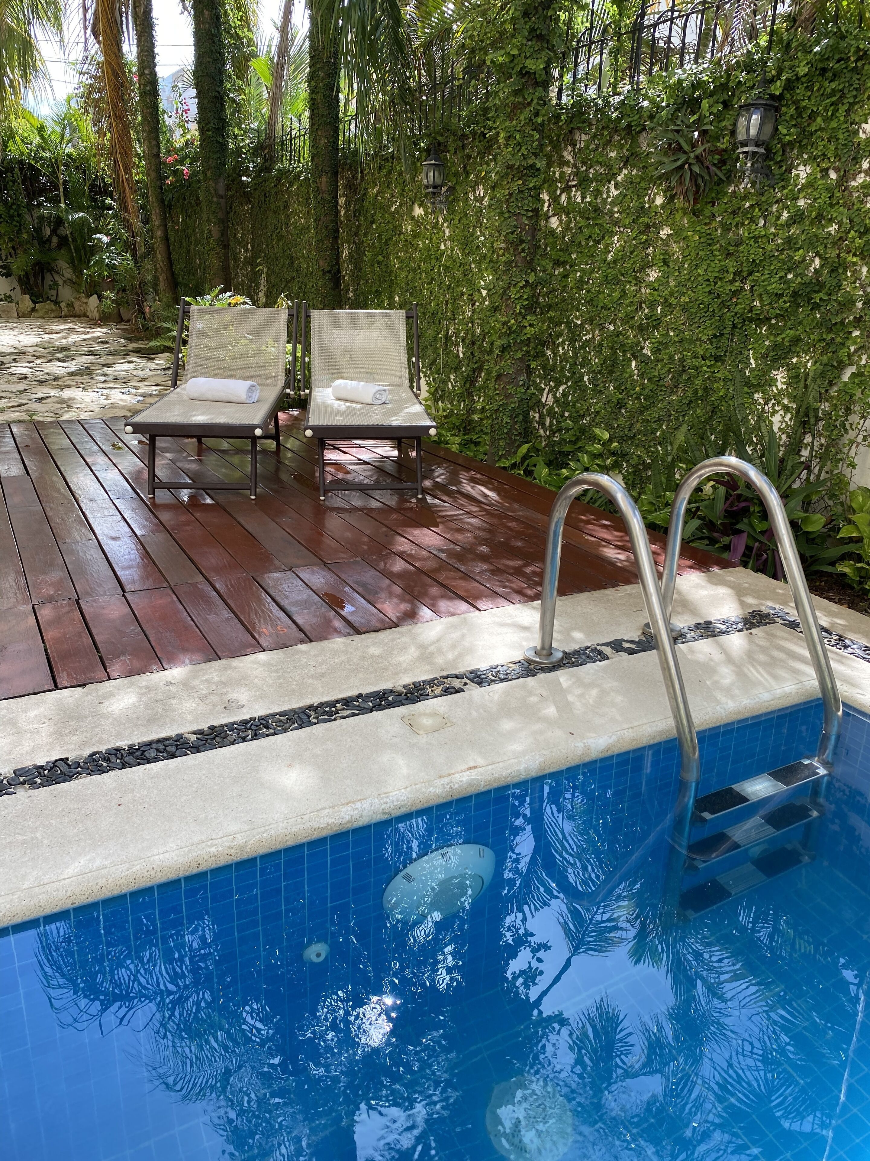 Un área de piscina con cubierta de madera, dos sillones de descanso y una escalera metálica para acceder al agua.