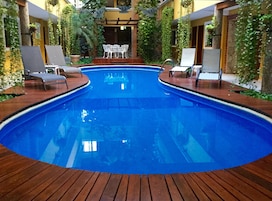 Piscina con sillones reclinables y una mesa en el centro, rodeada de follaje y una terraza de madera.
