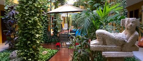 A courtyard with a wooden pathway, a stone statue, and lush greenery.