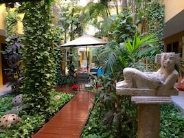 Un patio con un sendero de madera, una estatua de piedra y abundante vegetación.